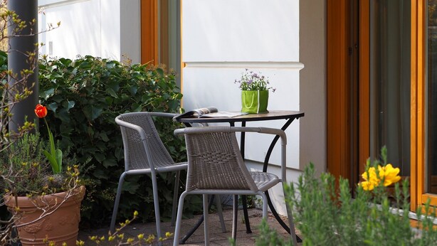 An outdoor seating area with wicker chairs and a table, surrounded by potted plants and a large window. - 1-room apartment with terrace (Seebad Heringsdorf)