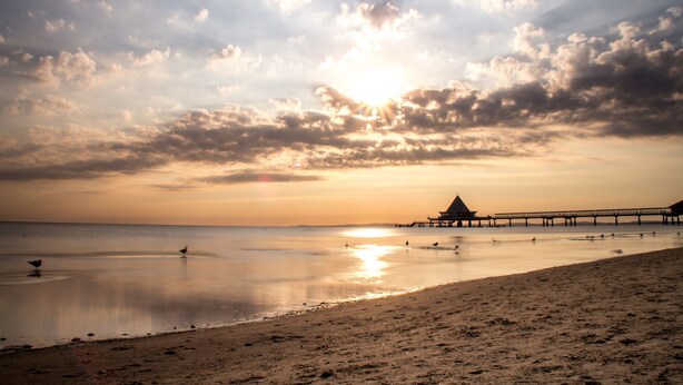 Une plage avec une jetée qui s’avance dans des eaux calmes, un ciel dégagé et un coucher de soleil.