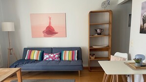 Un salon avec un canapé bleu, des coussins colorés, une bibliothèque en bois, une table blanche et un tapis gris.