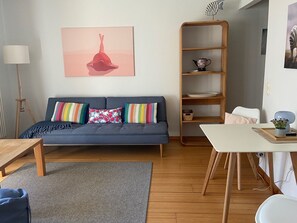 A living room with a blue sofa, colorful pillows, a wooden bookshelf, a white table, and a grey rug.