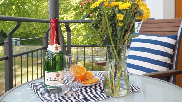 Une table avec une bouteille de Rothäppchen, deux verres, un vase de fleurs et des tranches d’orange, sur un balcon doté d’une balustrade et offrant une vue sur les arbres.
