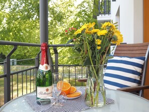 Une table avec une bouteille de Rothäppchen, deux verres, un vase de fleurs et des tranches d’orange, sur un balcon doté d’une balustrade et offrant une vue sur les arbres.
