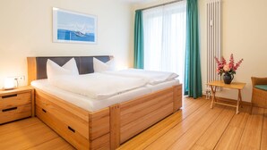 Camera d'albergo con un letto di legno, un comodino, una sedia, un tavolino con un vaso di fiori e una finestra con tende.