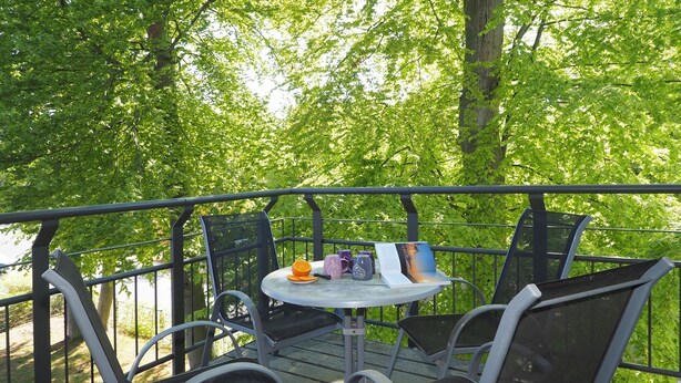 Terraço com cadeiras e mesa de metal, rodeado por grades metálicas, com vista para uma área arborizada.