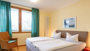 A hotel room with two beds, a wicker chair, a red nightstand, and a painting on the wall.