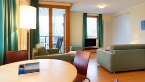 A modern hotel room with a round dining table, two sofas, a TV, and a balcony with curtains.