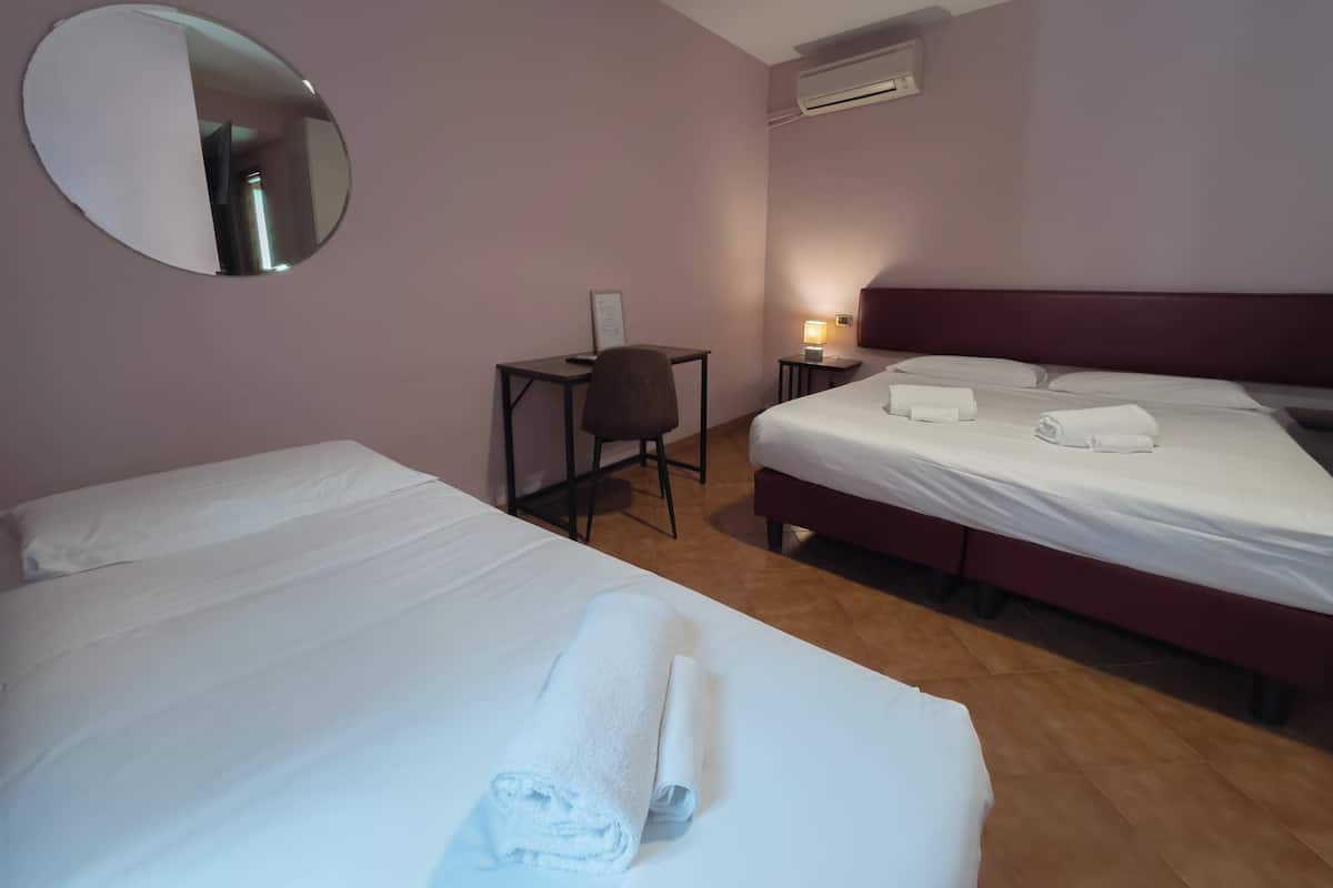 Habitación de hotel con dos camas, un escritorio, una silla, un espejo redondo, un equipo de aire acondicionado y una mesita de noche con lámpara.