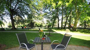 Uma área de estar externa com mesa e duas cadeiras, cercada por um parque com árvores e um caminho.