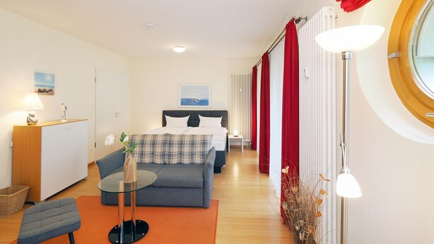 Quarto de hotel com cama, sofá, mesa pequena, luminária, janela com cortinas e radiador.
