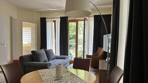 A modern living room with a sofa, a dining table with chairs, and a view of greenery outside. - 1-raum-appartement mit Balkon (Seebad Heringsdorf)