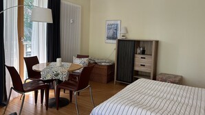 A hotel room with a bed, a dining table with chairs, a lamp, a radiator, and a window with curtains. - 1-raum-appartement mit Balkon (Seebad Heringsdorf)