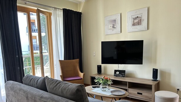 A living room with a sofa, a chair, a TV, speakers, and a small table with a vase. - 1-raum-appartement mit Balkon (Seebad Heringsdorf)