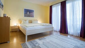 A bedroom with a bed, a nightstand, a wooden wardrobe, a painting on the wall, and a window with curtains. - Appartement mit 1 Schlafzimmer und Balkon (Seebad Heringsdorf)