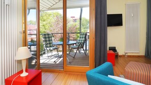 Ein Zimmer mit einer Schiebetür zu einem Außenbereich mit zwei Stühlen und einem Tisch. An der Wand ist ein Fernseher angebracht, es gibt einen roten Beistelltisch und ein blaues Sofa.