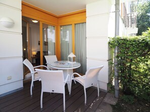 Een balkon met een ronde glazen tafel en vier rieten stoelen, een kleine witte lantaarn en uitzicht op een gebouw met een houten gevel.