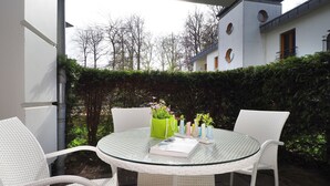 A round glass table with white wicker chairs, surrounded by potted plants, in an outdoor setting with a hedge and a building in the background. - Apartment with 1 bedroom and terrace (Seebad Heringsdorf)