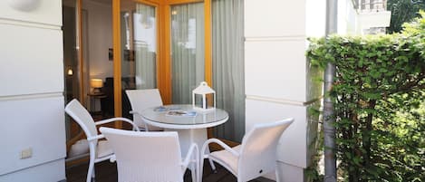 A balcony with a round glass table and white wicker chairs, a small lamp, and a view of a building with a wooden facade.