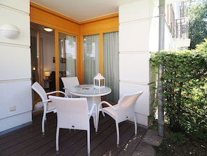 Ein Balkon mit rundem Glastisch und weißen Korbstühlen, einer kleinen Lampe und Blick auf ein Gebäude mit Holzfassade.