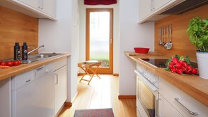 A narrow kitchen with wooden countertops, a dishwasher, and a stove. There is a window with a red curtain, a wooden chair, and a basket on the counter. - Apartment with 1 bedroom and terrace (Seebad Heringsdorf)