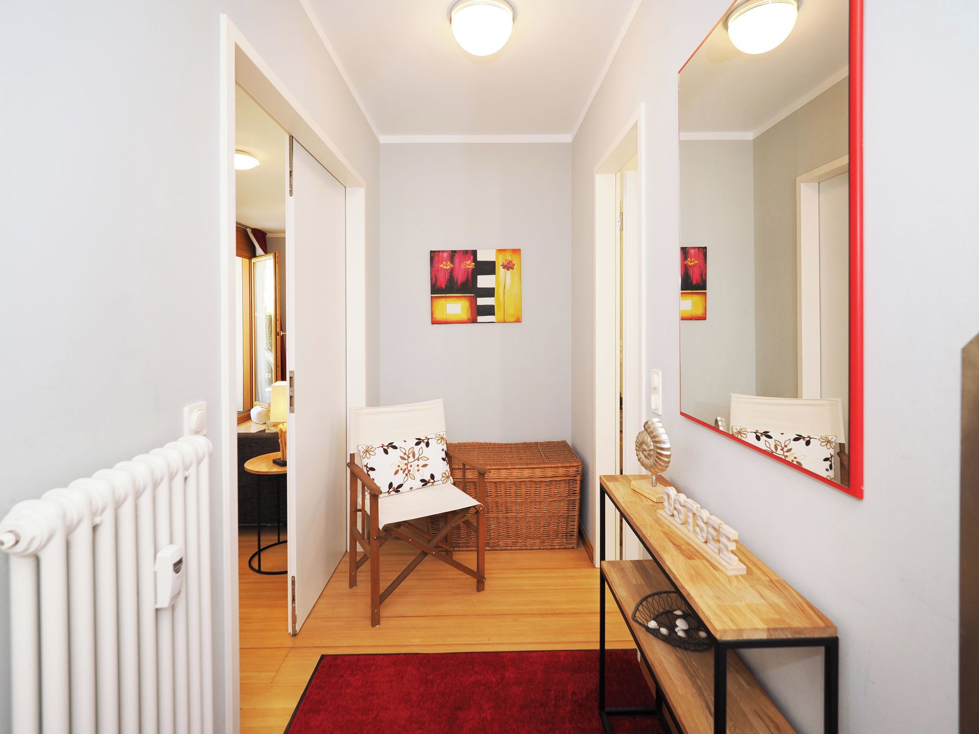 Ein Flur mit Holzboden, einem roten Teppich, einem weißen Heizkörper, einem Korbwäscher, einer Holzbank, einem Spiegel mit rotem Rahmen und zwei abstrakten Bildern an der Wand.