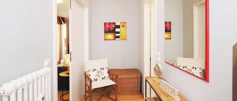 Ein Flur mit Holzboden, einem roten Teppich, einem weißen Heizkörper, einem Korbwäscher, einer Holzbank, einem Spiegel mit rotem Rahmen und zwei abstrakten Bildern an der Wand.