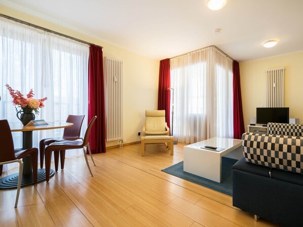 Ein modernes Hotelzimmer mit Essbereich, einer Couch, einem Fernseher und einem fensterbepflanzten Fenster.