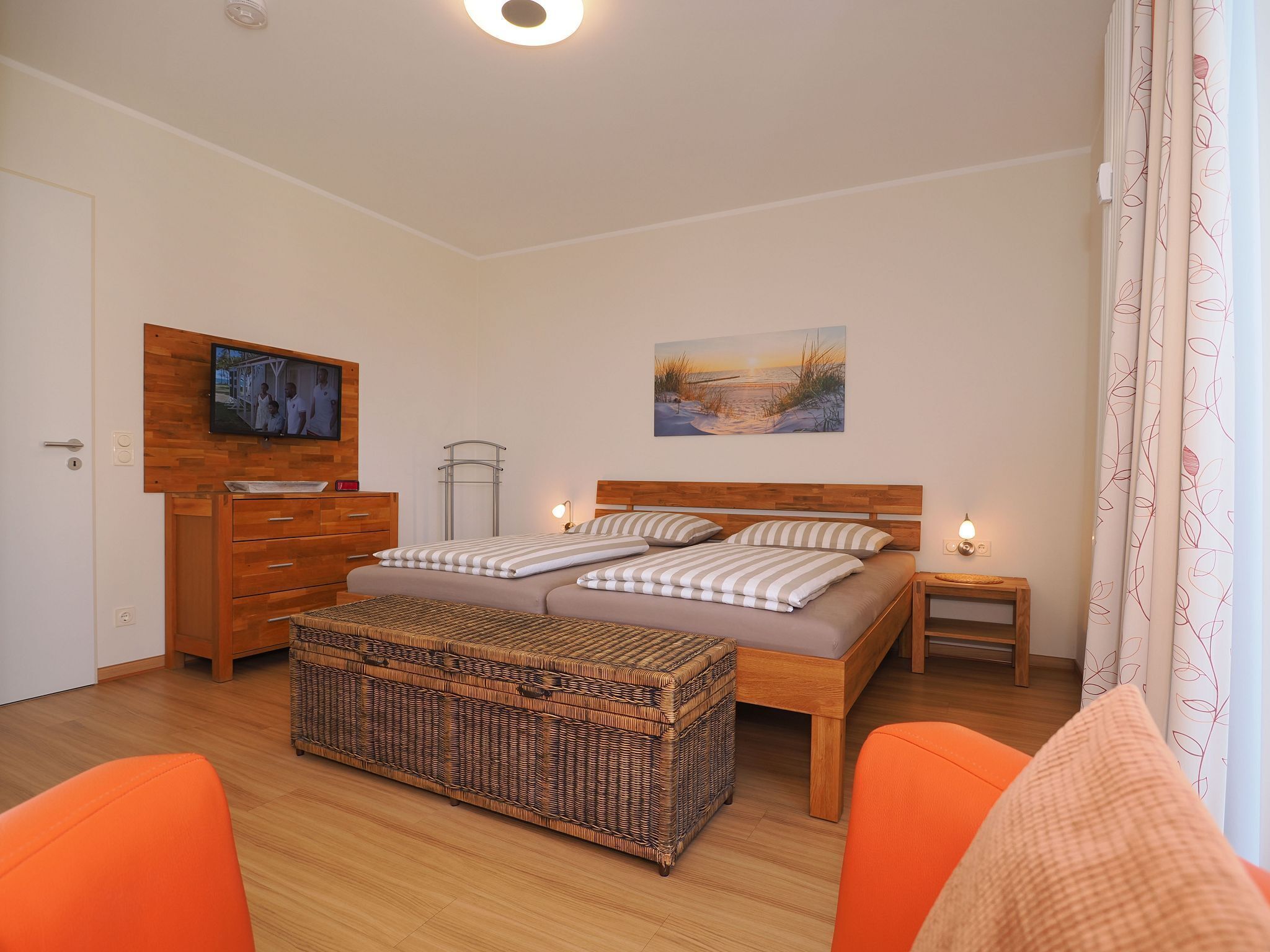Une chambre à coucher avec un lit, une télévision, un tableau, une petite table et une commode en osier.