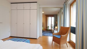 Et hotelværelse med en seng, en fletstol, et skrivebord med lampe og udsigt ud af store vinduer.
