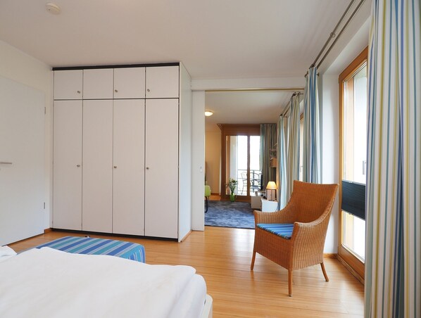 Hotellihuone, jossa on sänky, rottinkituoli, työpöytä lampun kanssa ja suuret ikkunat, joista on näkymä ulos.