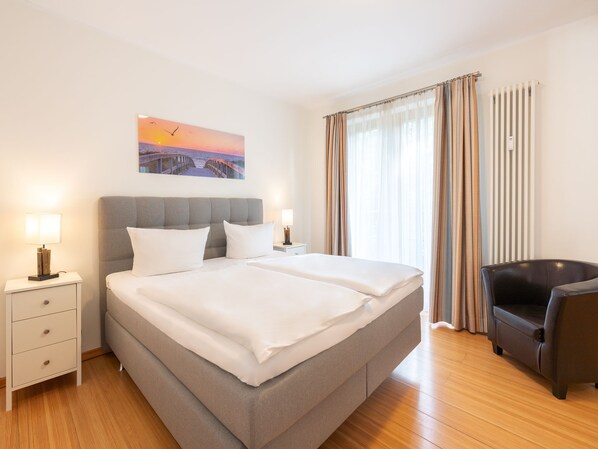 Une chambre d’hôtel avec un grand lit, deux tables de chevet, une chaise, un tableau représentant un coucher de soleil et une fenêtre avec des rideaux.
