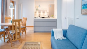 En moderne stue med en blå sofa, et træspisebord og stole. Der er et køkken i baggrunden med en køkkenø og hængende lamper.
