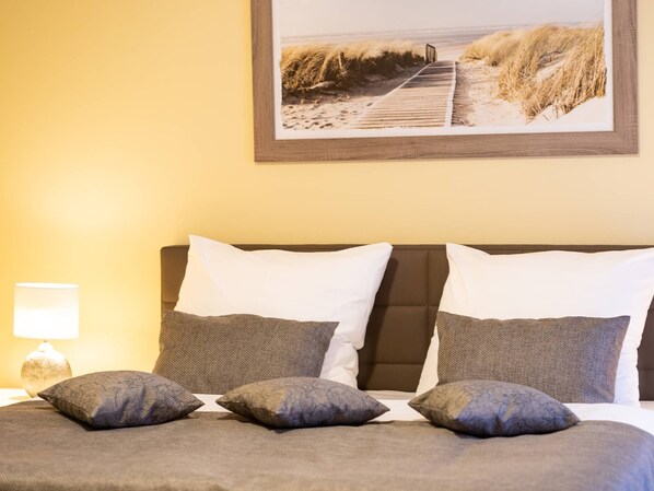 Quarto com cama, almofadas, mesa de cabeceira, abajur e um quadro na parede.