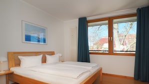 A neatly made bed with white linens in a room with wooden flooring, a window with blue curtains, and a painting on the wall. - Apartment with 1 bedroom and balcony (Seebad Heringsdorf)
