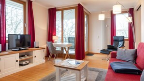 A modern living room with a flat-screen TV, a dining table, a sofa, and a red couch. - Apartment with 1 bedroom and balcony (Seebad Heringsdorf)