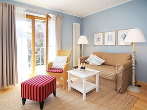 Een gezellige woonkamer met een bank, een rieten fauteuil, een gestreept voetenbankje, een kleine tafel met flessen en lijstwerken aan de muur.