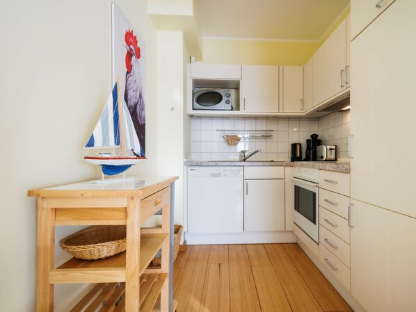 A compact kitchen with white cabinets, a microwave, a sink, and a wooden table with a sailboat model. - 1-room apartment with balcony (Seebad Heringsdorf)