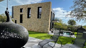 Un espacio exterior moderno con un edificio de piedra, una escultura negra decorativa y una piscina con sillas de descanso.