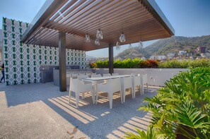 An outdoor patio with white tables and chairs, a wooden pergola, and hanging lights.