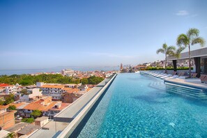 A large outdoor swimming pool with clear blue water, surrounded by lounge chairs and palm trees, overlooking a coastal town with buildings and a distant city skyline.