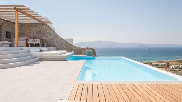 Área de piscina moderna ao ar livre, com muro de pedra, deck de madeira e vista para o mar.