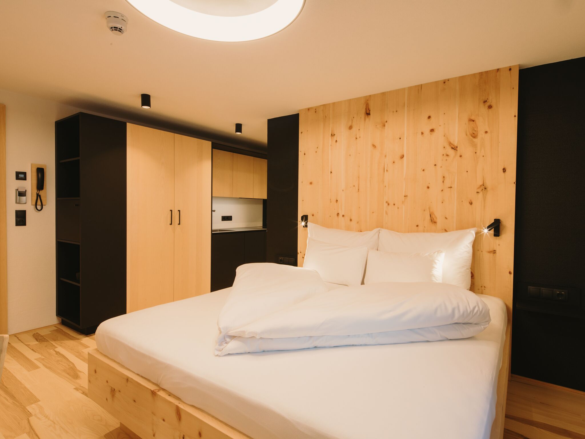 A modern bedroom with a wooden headboard, a large bed, built-in wardrobe, and a small kitchenette.
