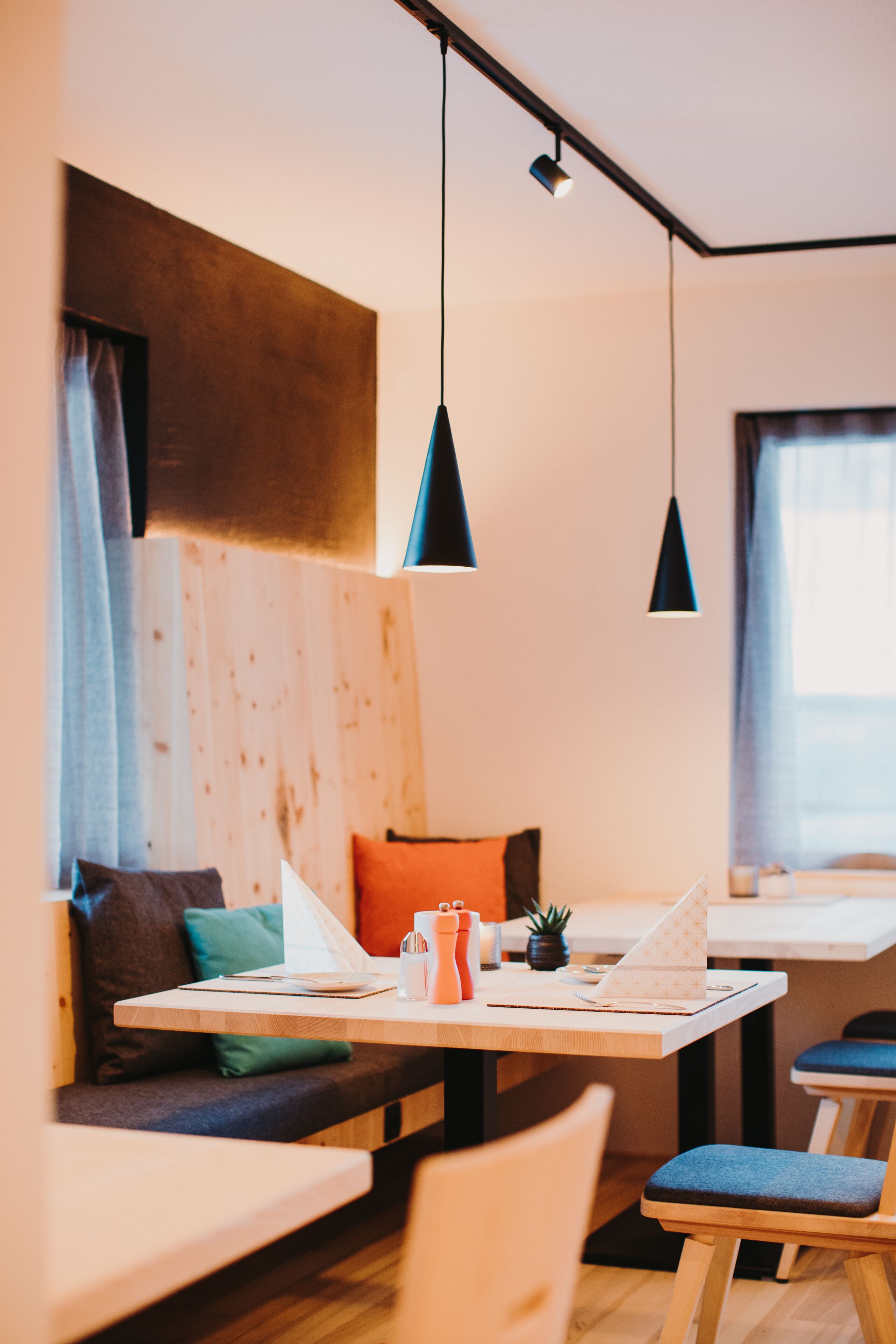 A modern interior with a wooden accent wall, a table with a laptop, a kettle, and a small plant. There are two hanging pendant lights and a window with curtains.