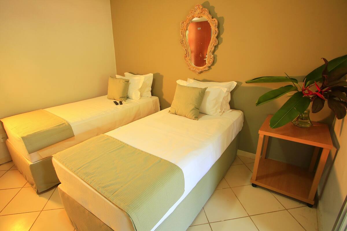 Habitación de hotel con dos camas individuales, una mesita de noche de madera con una planta y un espejo en la pared.