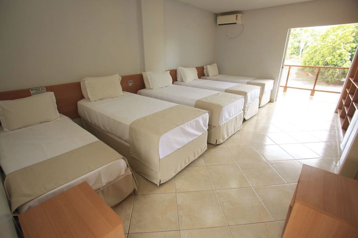 Habitación de hotel con cuatro camas individuales alineadas, cabecera de madera, suelo de baldosas y una puerta corrediza de vidrio que da a un espacio exterior.