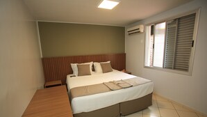 Une chambre à coucher avec un lit, une table de chevet en bois, une fenêtre avec des stores et un sol carrelé.