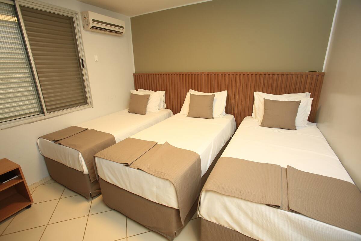 Habitación de hotel con tres camas individuales, cabecero de madera, ventana con persianas y unidad de aire acondicionado.