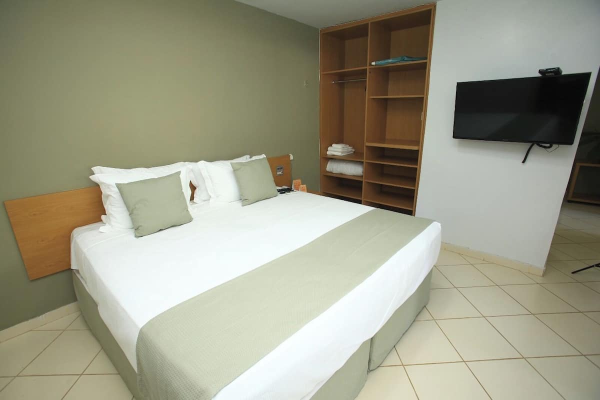 Una cama bien hecha con cabecera de madera, un televisor montado en la pared y un amplio armario con estantes.
