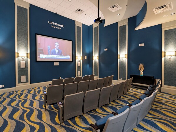Un salón de conferencias con una pantalla grande mostrando un noticiero, filas de sillas y una alfombra estampada.
