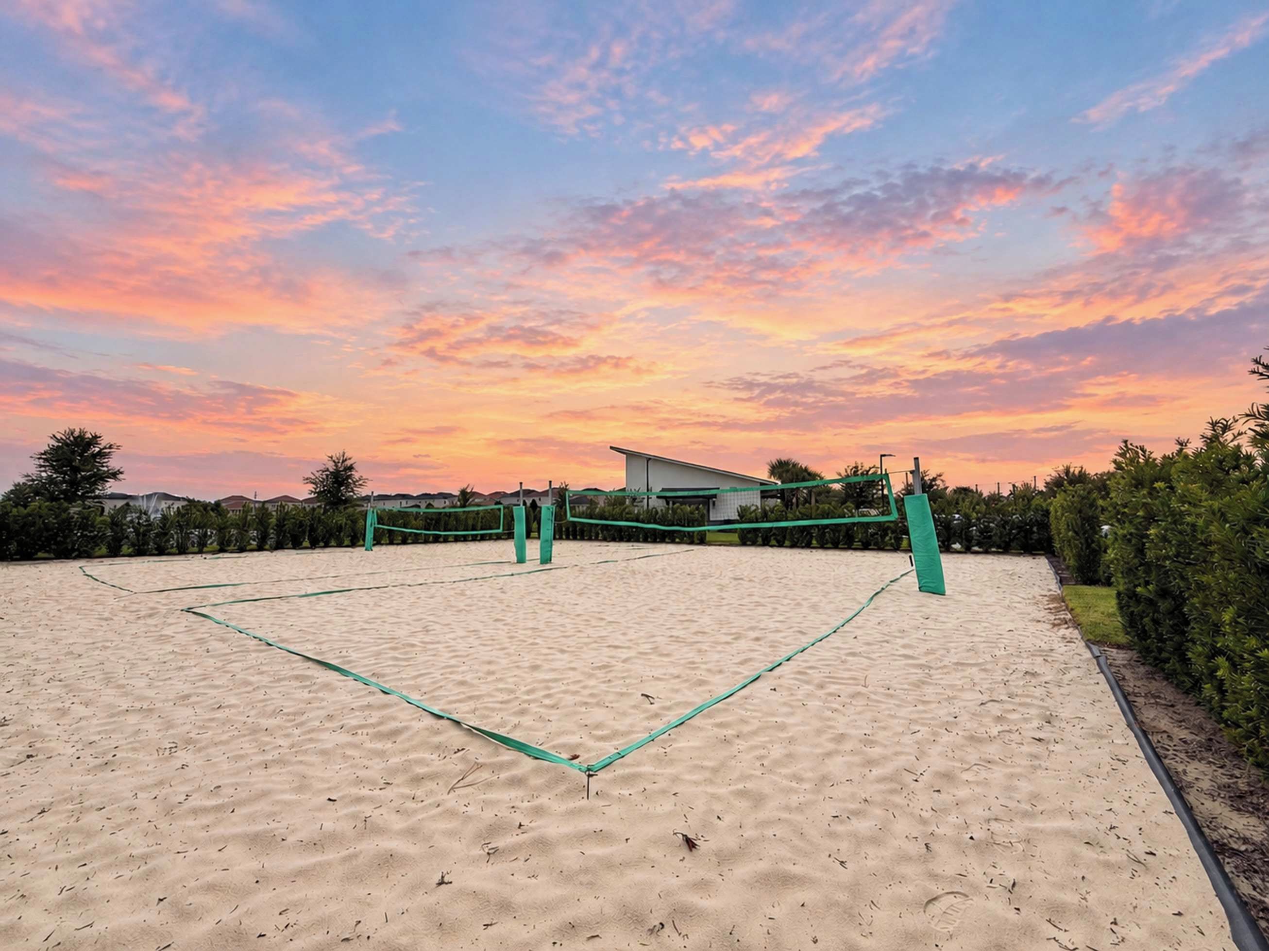 A volleyball court with a net, surrounded by sand and greenery, under a sunset sky.
