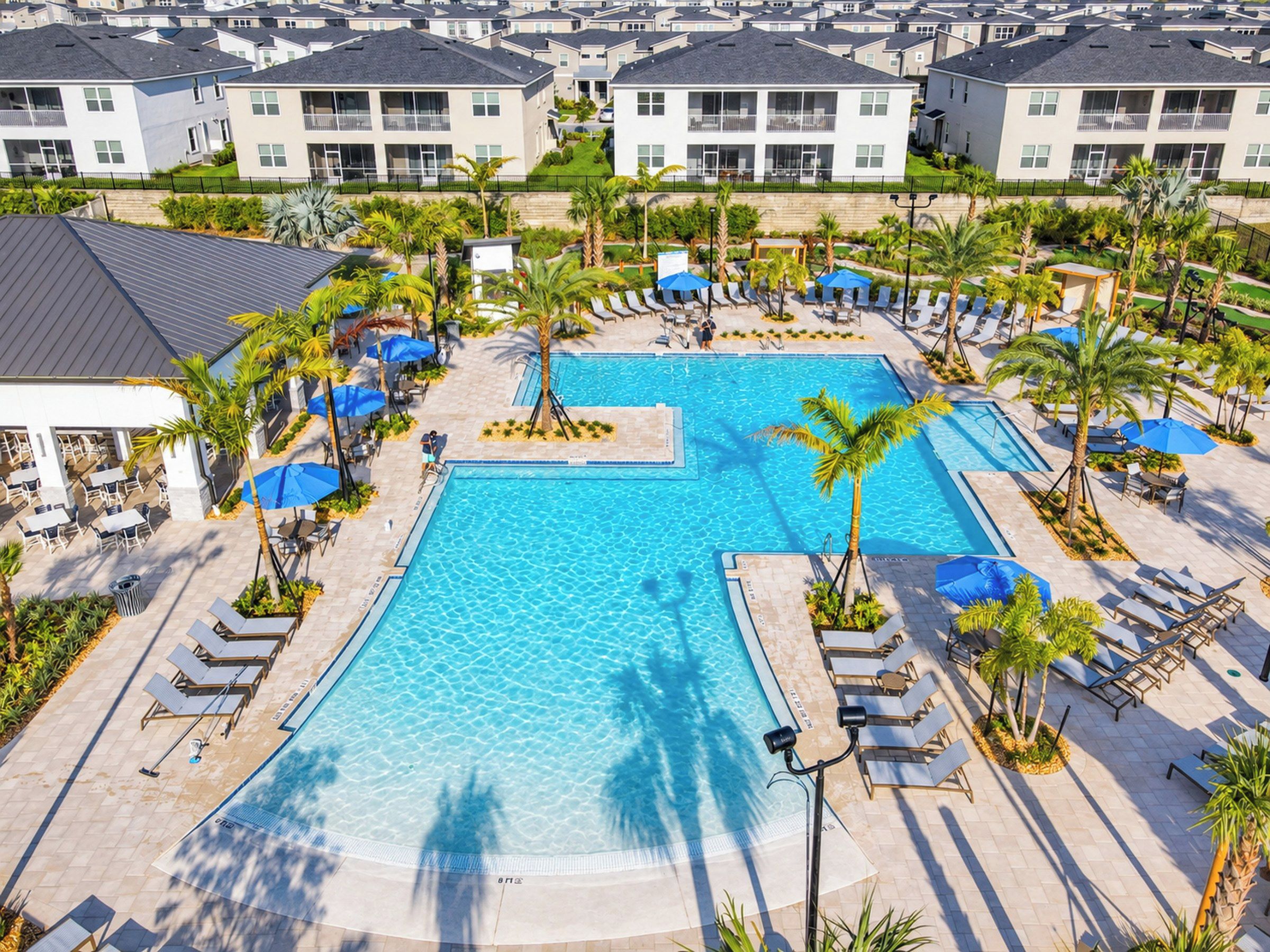 A large swimming pool surrounded by lounge chairs and umbrellas, with a view of residential buildings in the background.